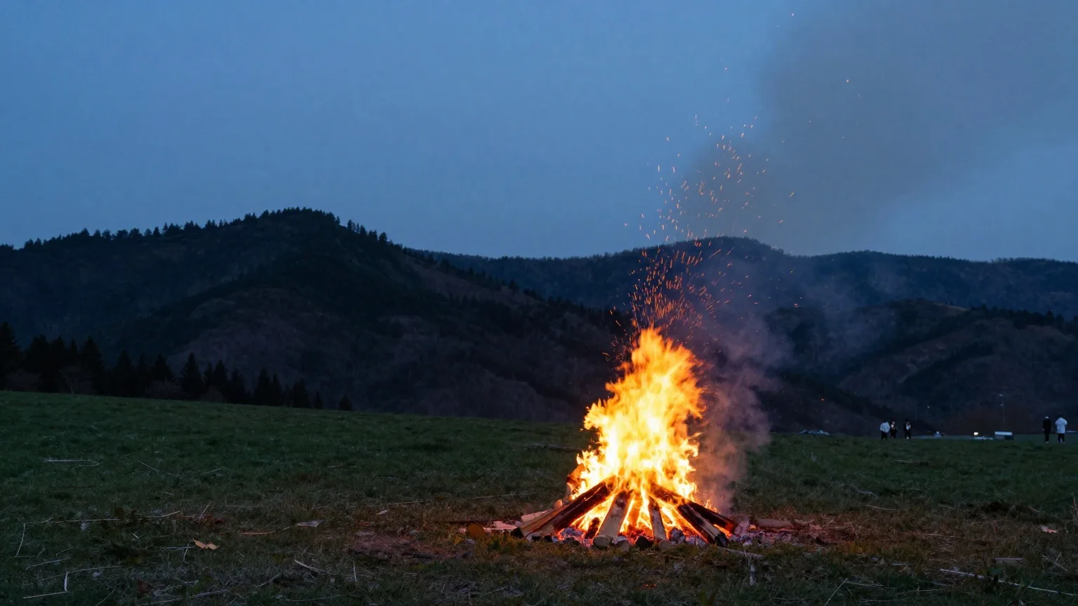 Lagerfeuer auf einer Bergwiese in der Abenddämmerung, dahinter bewaldete Hügel