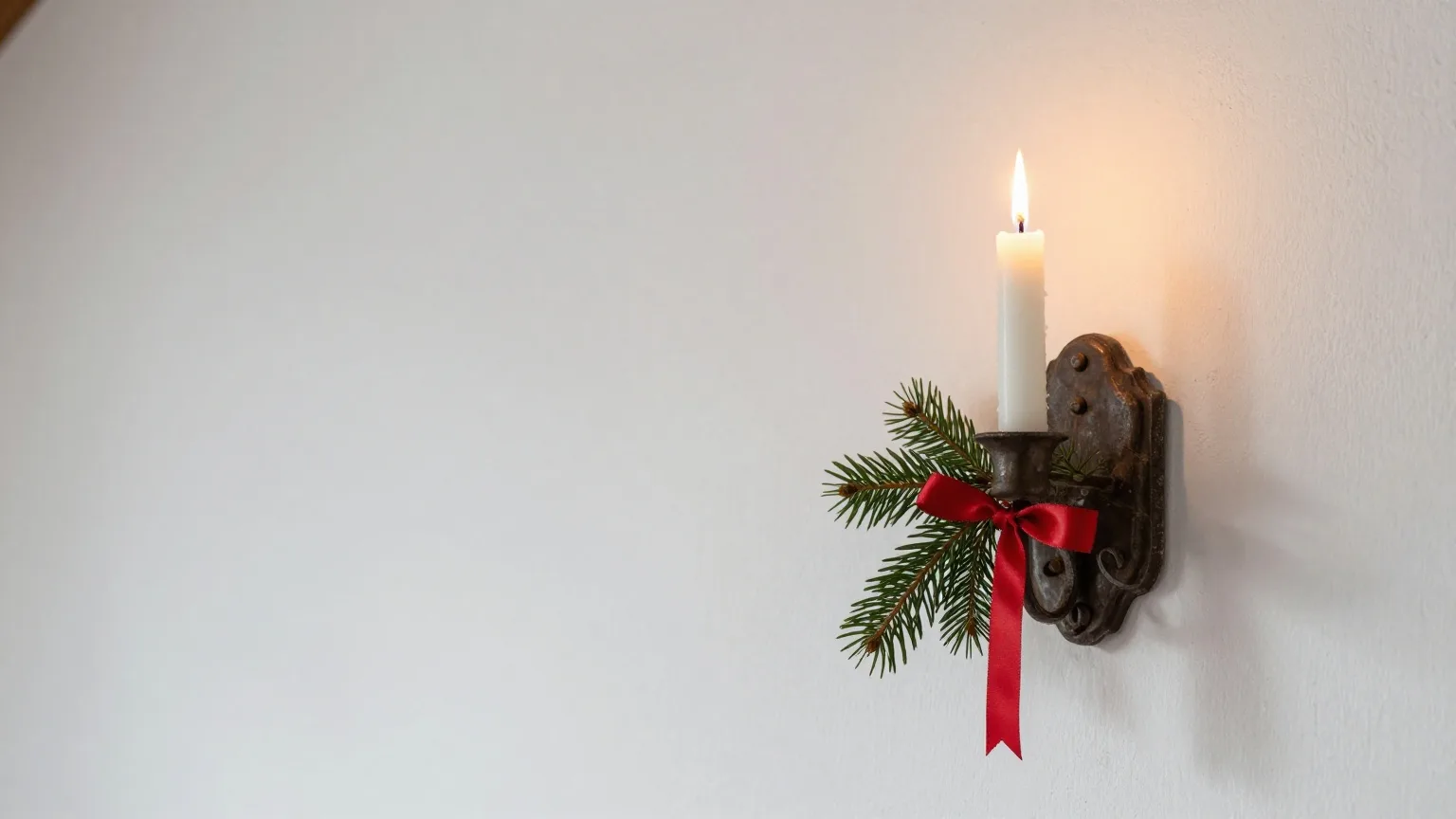 Einzelne brennende Kerze in einem schmiedeeisernen Wandleuchter an einer weiß gekalkten Wand, daneben ein Tannenzweig mit roter Schleife