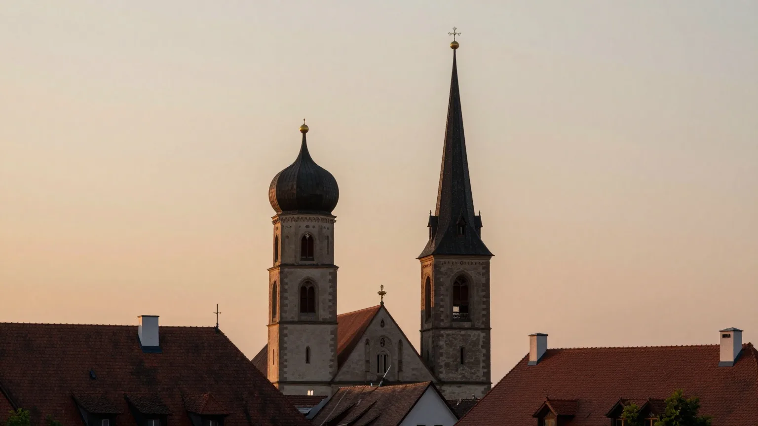 Silhouetten zweier unterschiedlich geformter Kirchtürme über roten Ziegeldächern im Abendlicht