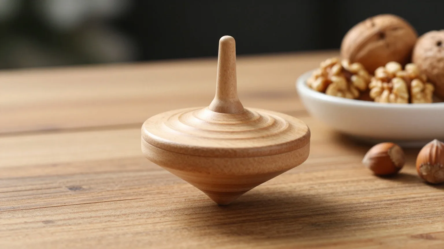 Hölzerner Kreisel auf einem Holztisch neben einer Schale Walnüsse, davor zwei Haselnüsse im Lampenlicht