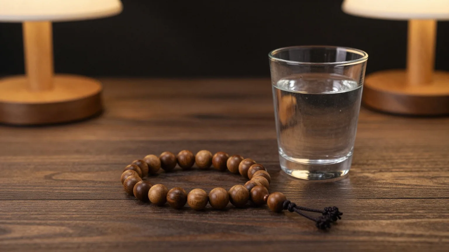 Gebetskette aus Olivenholzperlen und ein Glas Wasser auf einem dunklen Holztisch im warmen Lampenlicht am Abend