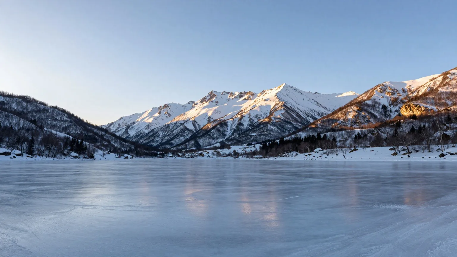 Zugefrorener Bergsee vor verschneiten Gipfeln im Licht der tief stehenden Nachmittagssonne