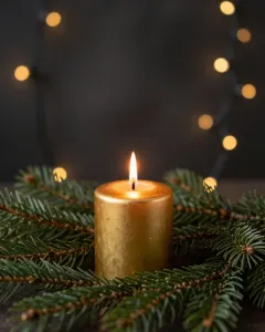 Eine brennende goldene Kerze zwischen Tannenzweigen vor warmen, unscharfen Lichtern im dunklen Raum