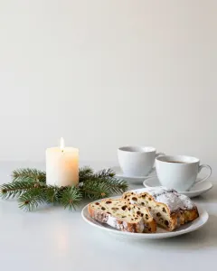 Eine brennende weiße Kerze in einem kleinen Tannenkranz neben Christstollen und zwei Kaffeetassen
