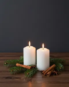 Zwei brennende weiße Stumpenkerzen auf Tannenzweigen mit Zimtstangen und Sternanis auf einem dunklen Holztisch