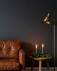 Zwei brennende grüne Kerzen auf Tannenzweigen neben einem Ledersessel im warmen Licht einer Stehlampe