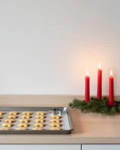 Drei brennende rote Kerzen auf Tannenzweigen neben einem Blech mit frisch gebackenen Sternplätzchen