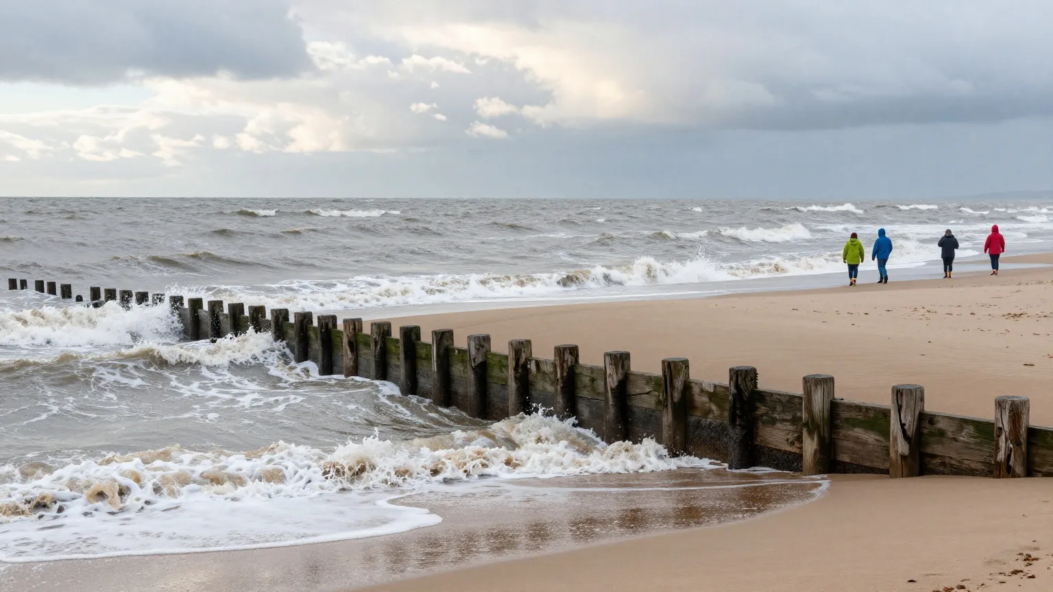 Hölzerne Buhnen am Nordseestrand bei Herbststurm, eine Familie in Regenjacken von hinten in der Ferne
