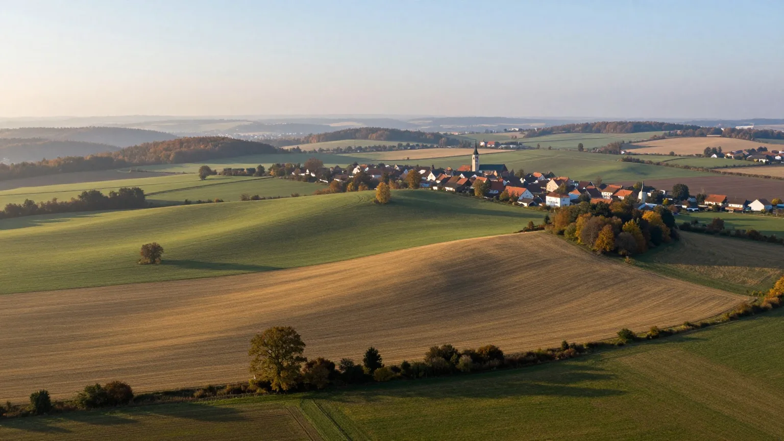 Hügelige Kulturlandschaft mit Feldern, Wäldern und einem Dorf mit Kirchturm aus der Luft