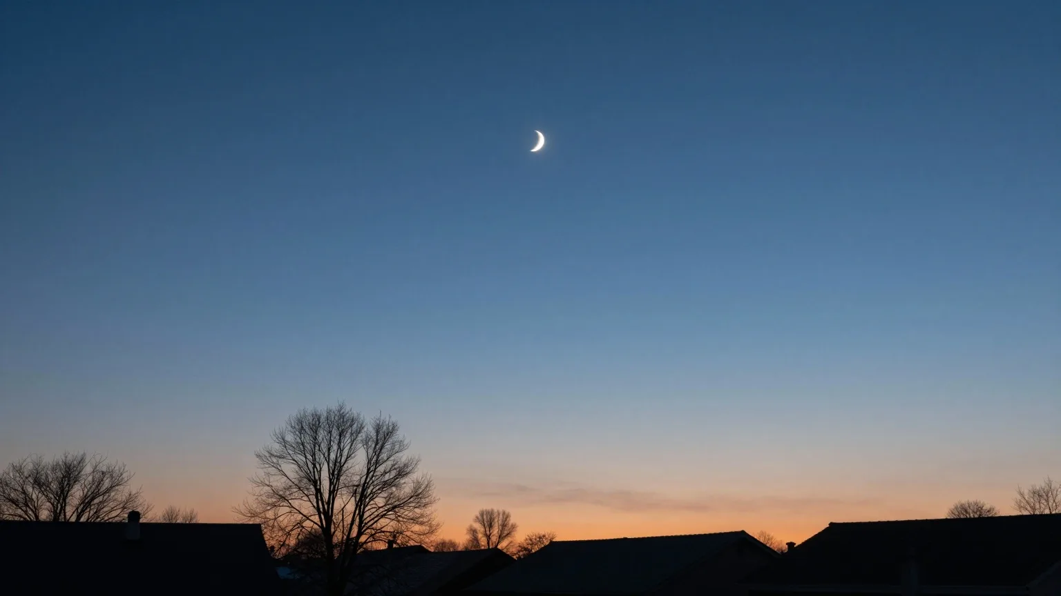 Schmale Mondsichel am dunkelblauen Abendhimmel über den Dächern eines Dorfes